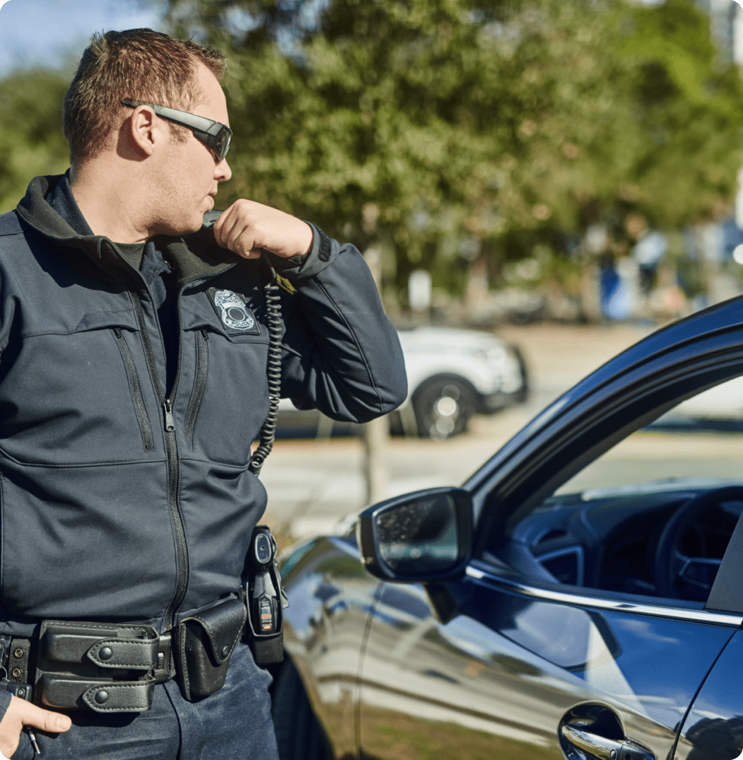 A police officer stands beside a car, speaking into a radio microphone, during a daytime traffic stop.