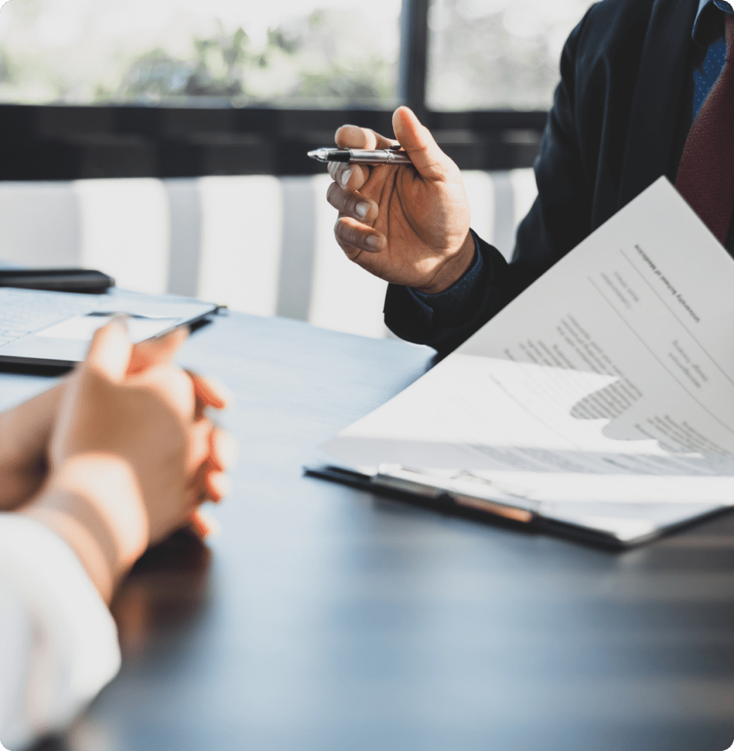 Two people sit across a table in a business meeting, with documents and a pen in hand, discussing charts and financial data.
