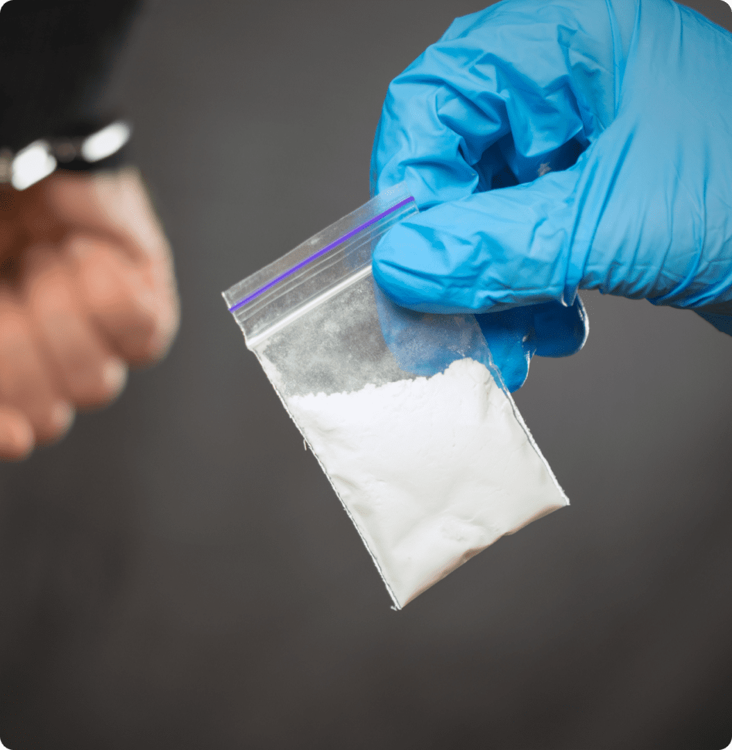 A gloved hand holds a small clear plastic bag containing white powder, while another person's cuffed hands are visible in the background.