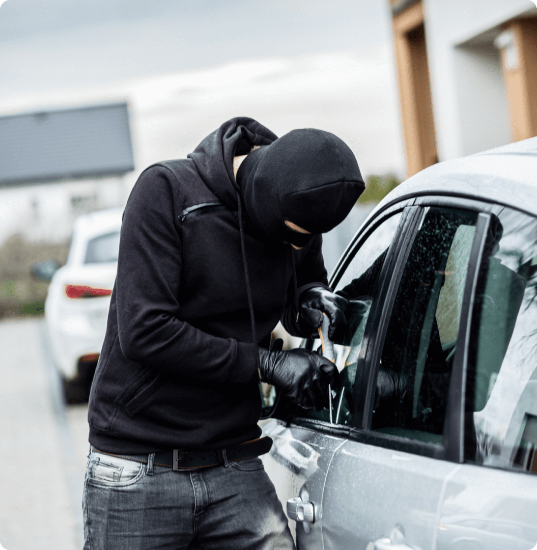 A person wearing a black balaclava and gloves attempts to break into a parked car using a tool.