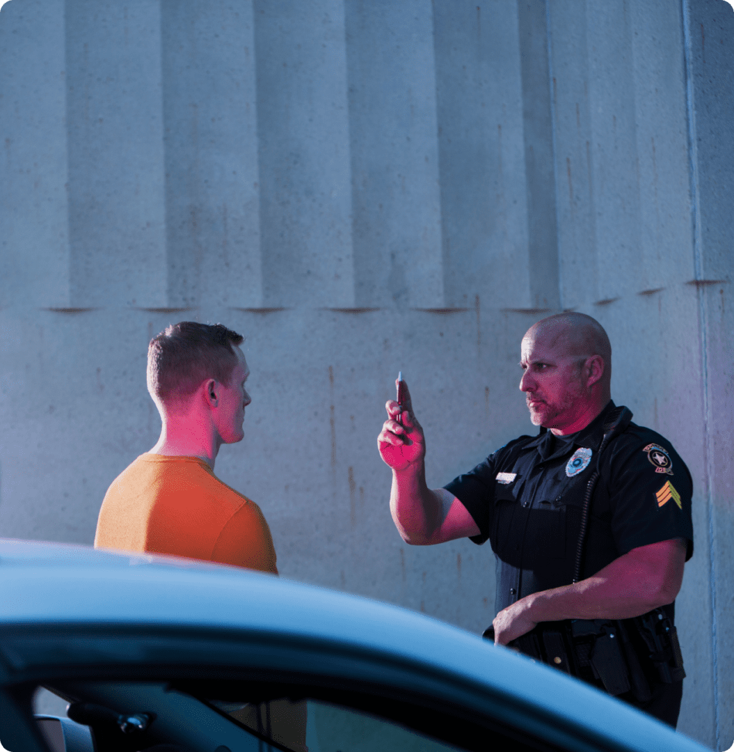 A police officer performs a sobriety test on a man by holding up a finger in front of him near a parked vehicle at dusk.