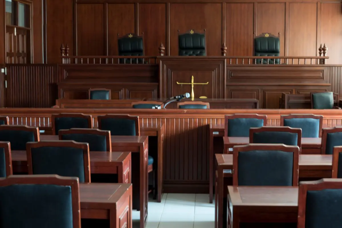 Empty courtroom with wooden desks and chairs, a judge’s bench, and a scale of justice symbol at the front.
