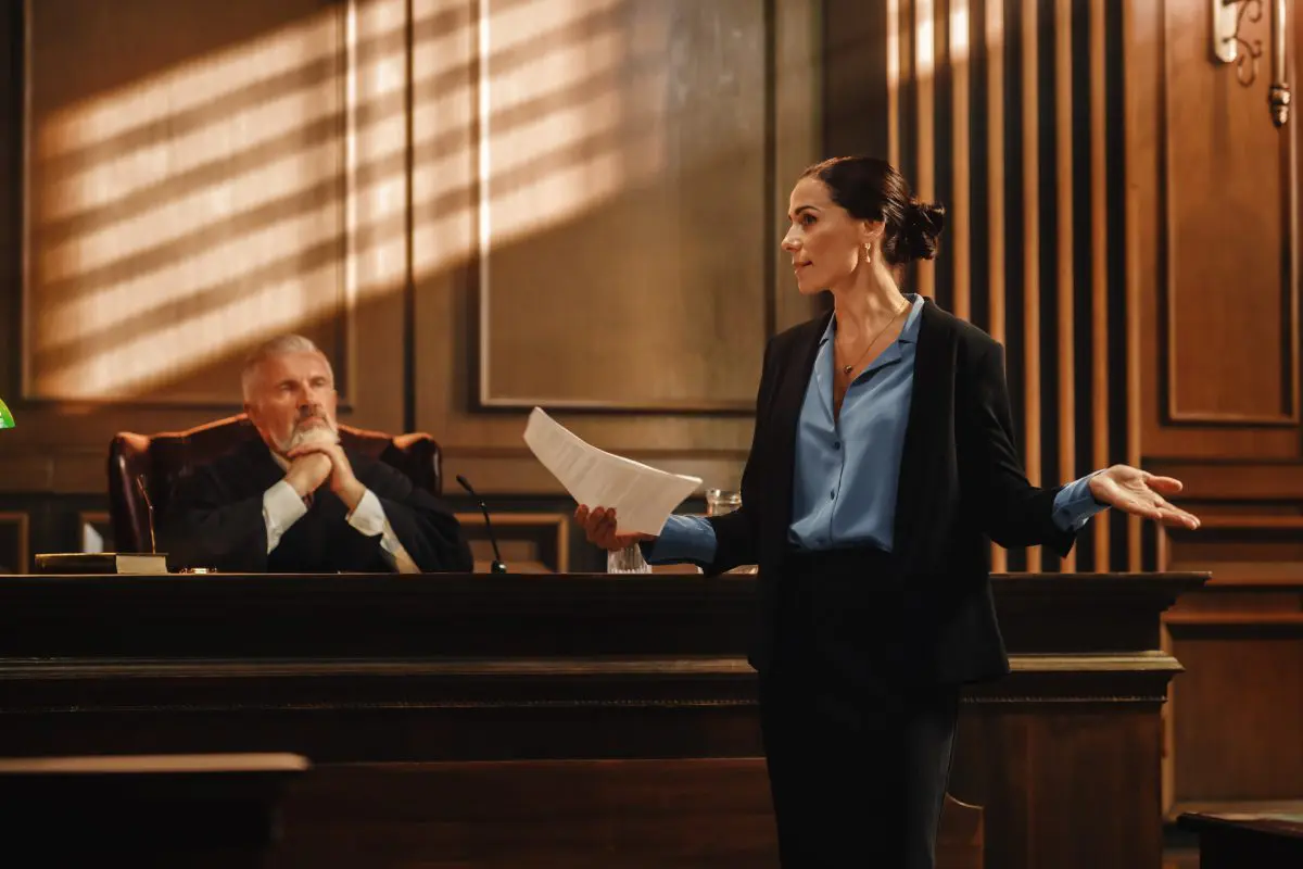 A female lawyer speaks while holding papers in a courtroom, as a judge sits and listens behind the bench.
